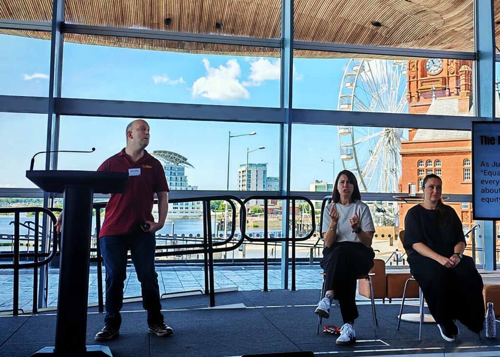 Gavin Clifton standing on a stage at the Welsh Senedd, delivering a talk.  In the background you can see Cardiff Bay