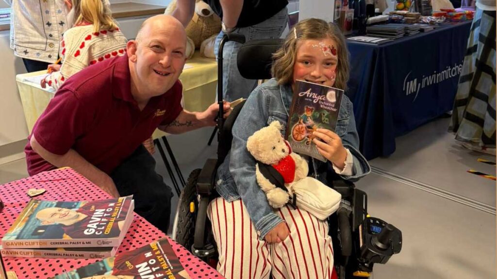 Gavin Clifton pictured with a young lady, Sienna who is seated in a wheelchair and holding a copy of Anya and the enchanted Wheelchair